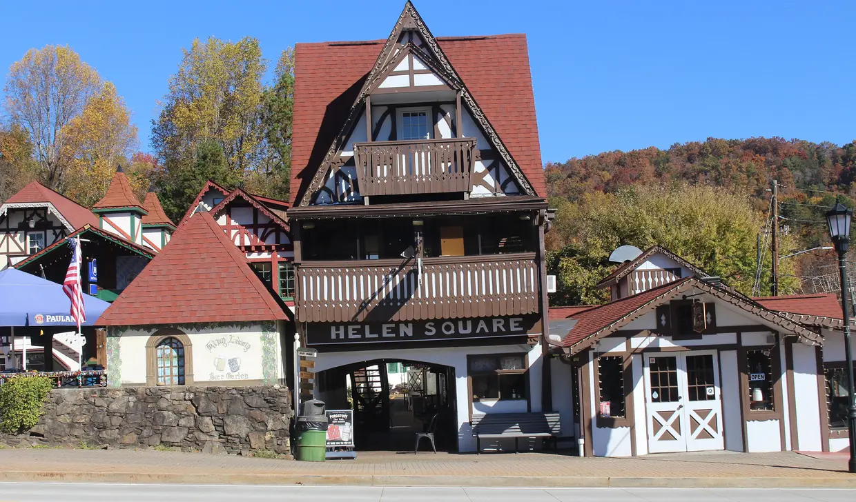 A tall half-timbered building with a carved balcony and a steep red roof above the arched entrance to Helen Square, with autumn trees and hills behind.