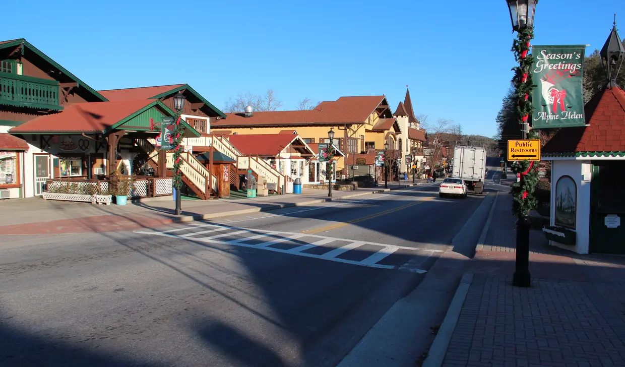 Main Street in Helen in winter sun: Bavarian-style shopfronts, and a street lamp wrapped in garland with a red "Season's Greetings" banner.
