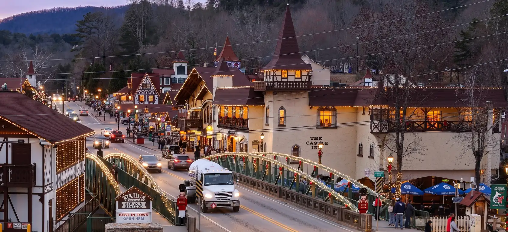 Evening illustration of downtown Helen: the arched bridge over the Chattahoochee River strung with white lights, The Castle Inn and half-timbered shops glowing along Main Street, wooded ridges behind.